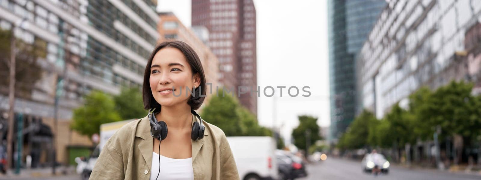 Portrait of young asian woman in headphones, posing in city, smiling and looking away, standing on street of city centre by Benzoix