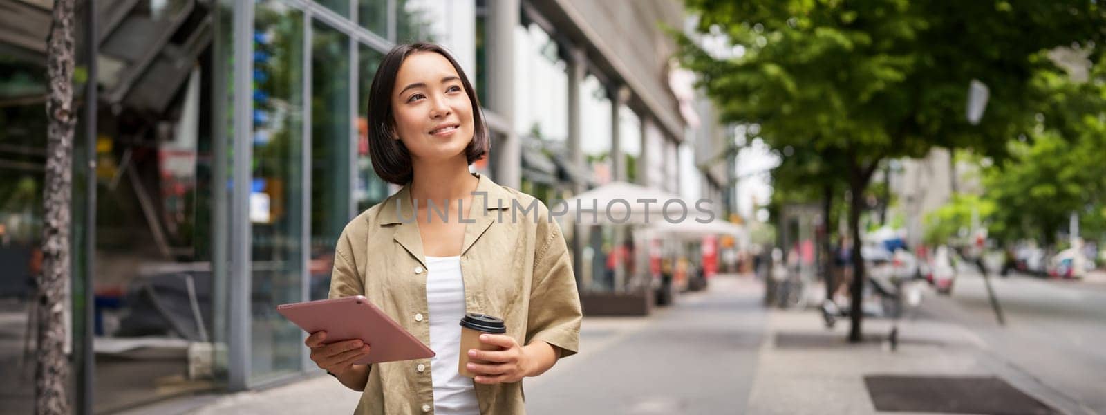 Portrait of smiling young woman walking in city with tablet, drinking takeaway coffee, going down the street with happy expression.