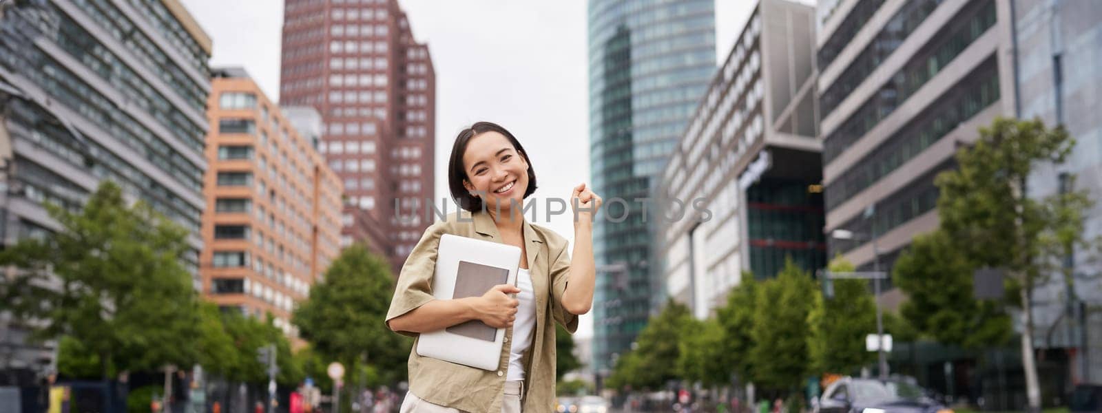 Portrait of happy asian woman stands with tablet near street road, cheering, raising hand up in triumph, celebrating by Benzoix