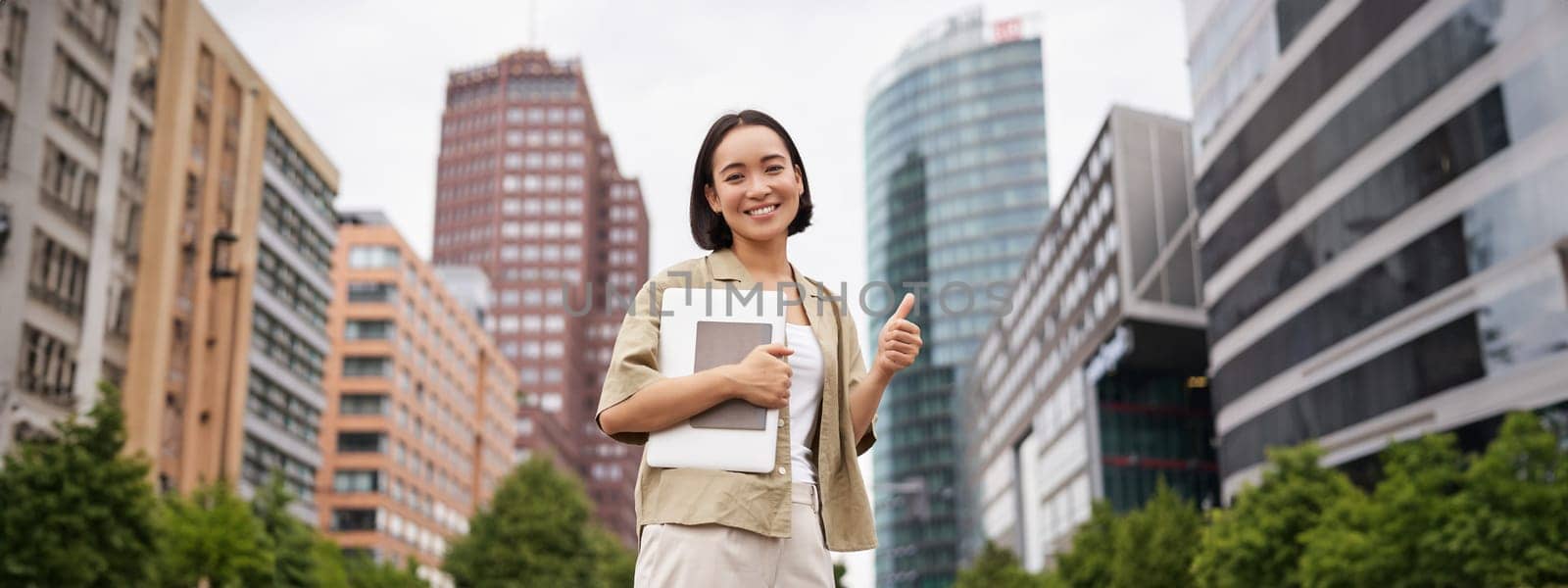 Cheerful asian girl in city centre, showing yes, hooray gesture, standing with laptop and celebrating.