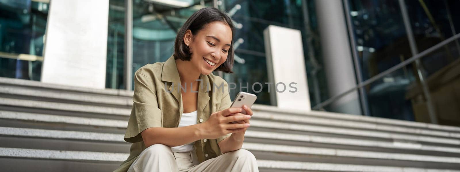 Technology and communication. Young smiling girl, asian woman sits with smartphone, reads message with big smile.