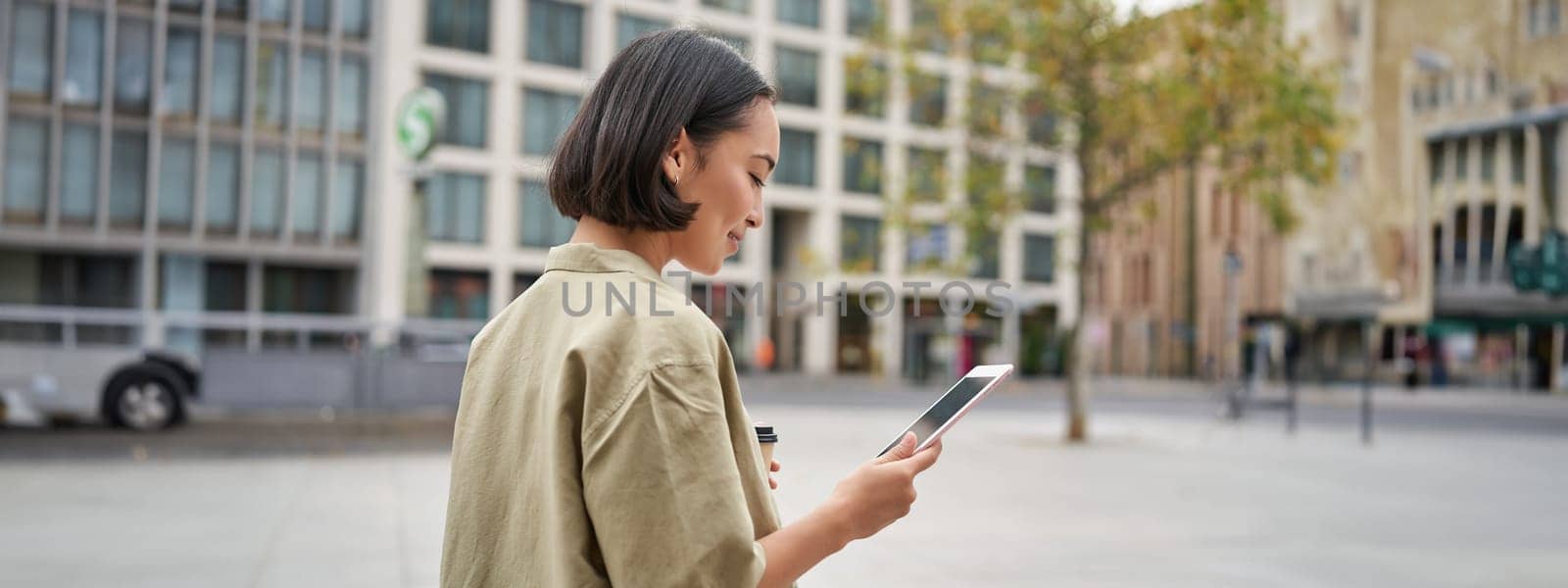 Portrait of stylish urban girl walks in city with tablet, drinks takeaway coffee.