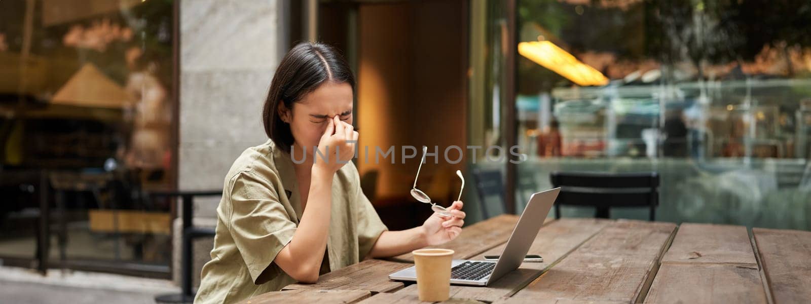 Young asian woman feeling tired after working with laptop, sitting in cafe on bench outdoors, drinking coffee, looking exhausted.