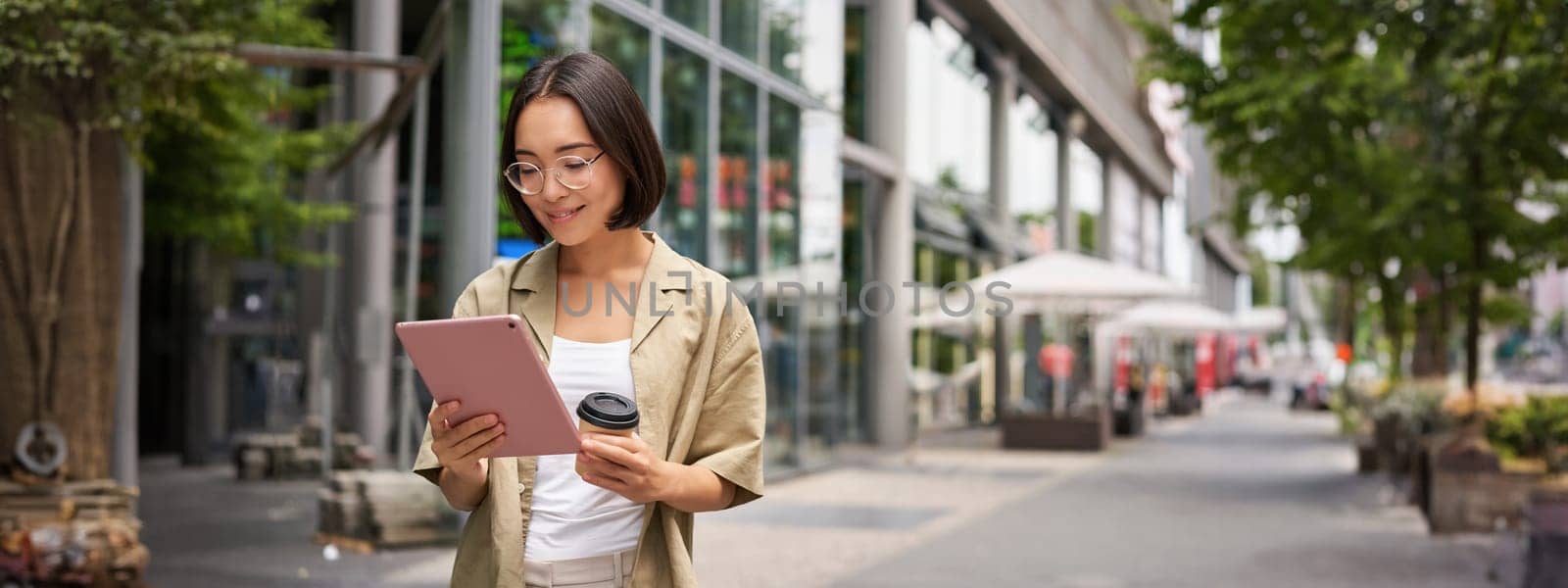 Portrait of smiling young woman walking in city with tablet, drinking takeaway coffee, going down the street with happy expression.