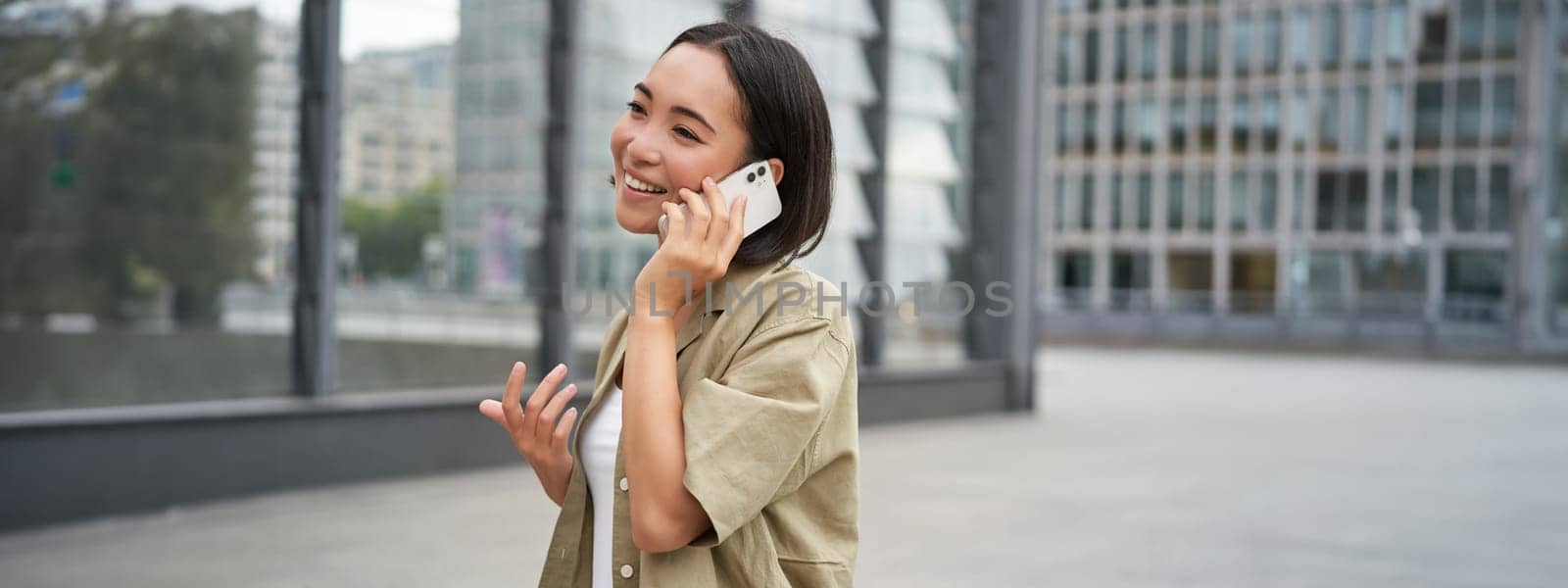 Cellular connection. Young asian woman makes a telephone call, talking on mobile smartphone and walking on street by Benzoix