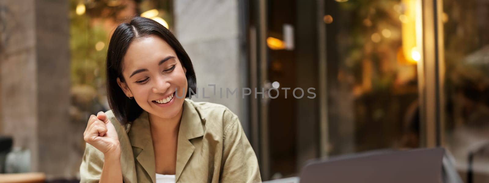 Vertical shot of happy girl talking on video call, looks at laptop, having online meeting, sitting in outdoor cafe.