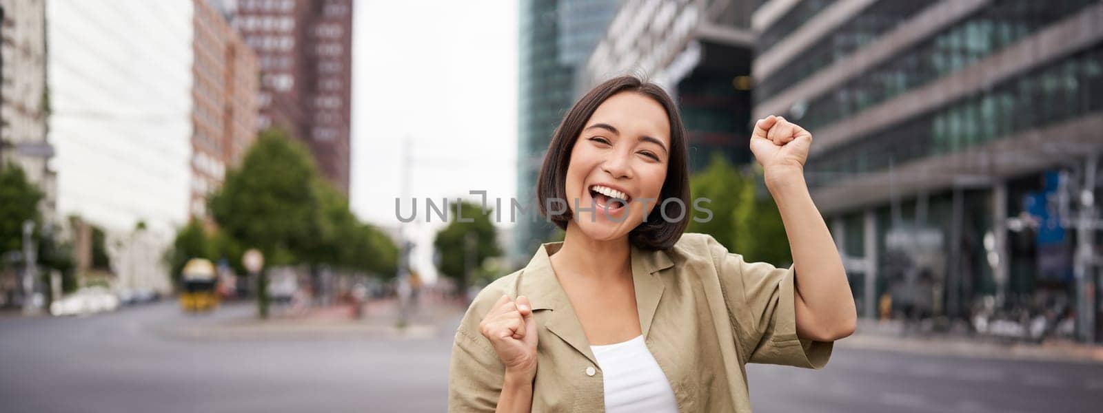 Happy asian girl winning and rejoicing, celebrating on streets of city, dancing from joy, triumphing and smiling.