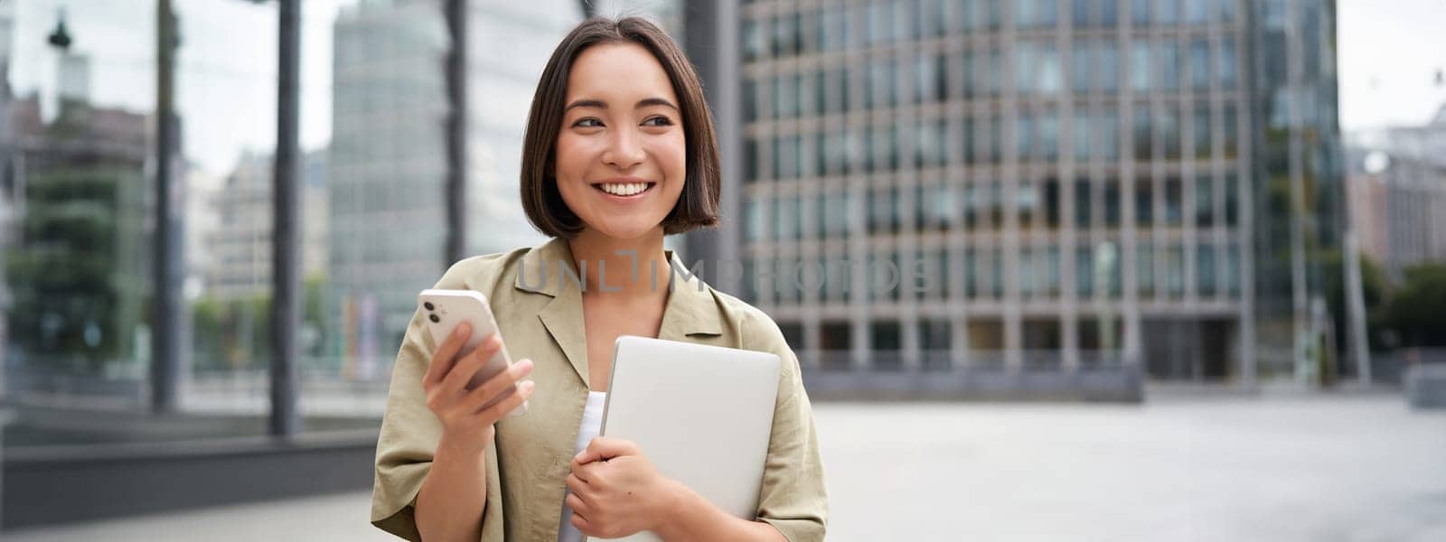 Portrait of young smiling asian woman walking on street, going to work with laptop and smartphone by Benzoix