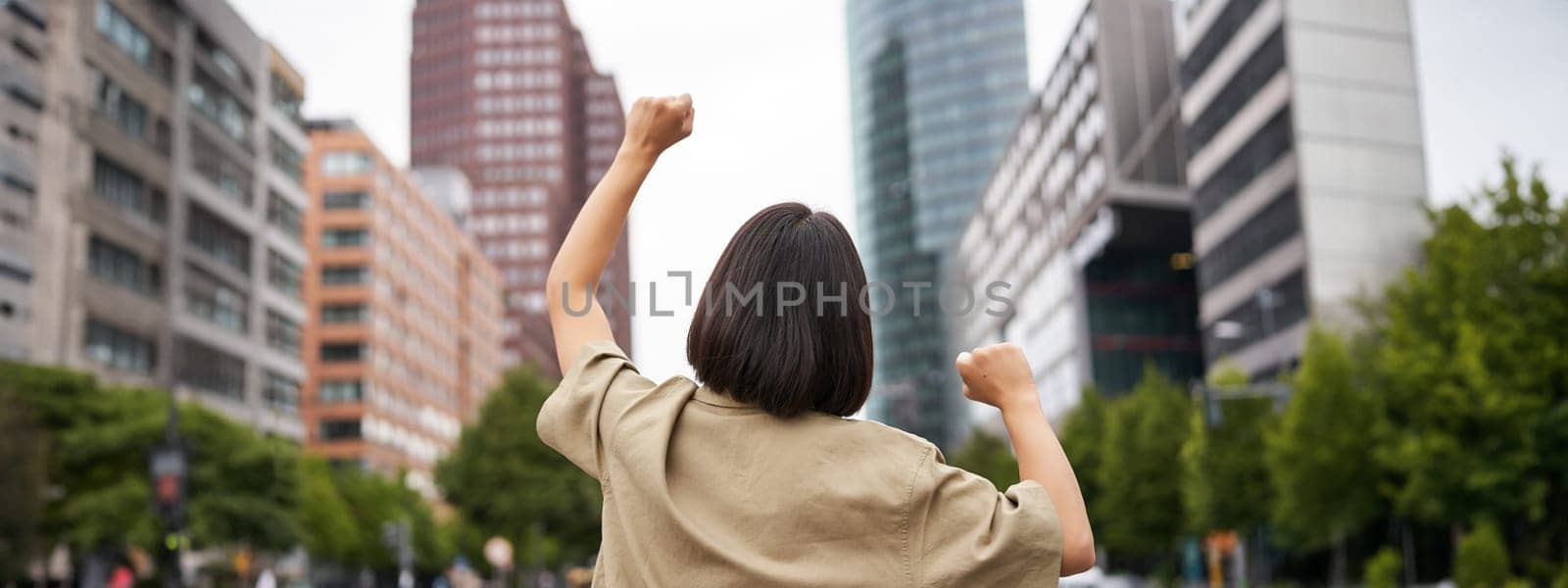 Excitement. Happy girl rear view, raising her hands up and looking at big city skyscrappers, feeling confident, cheering and triumphing by Benzoix