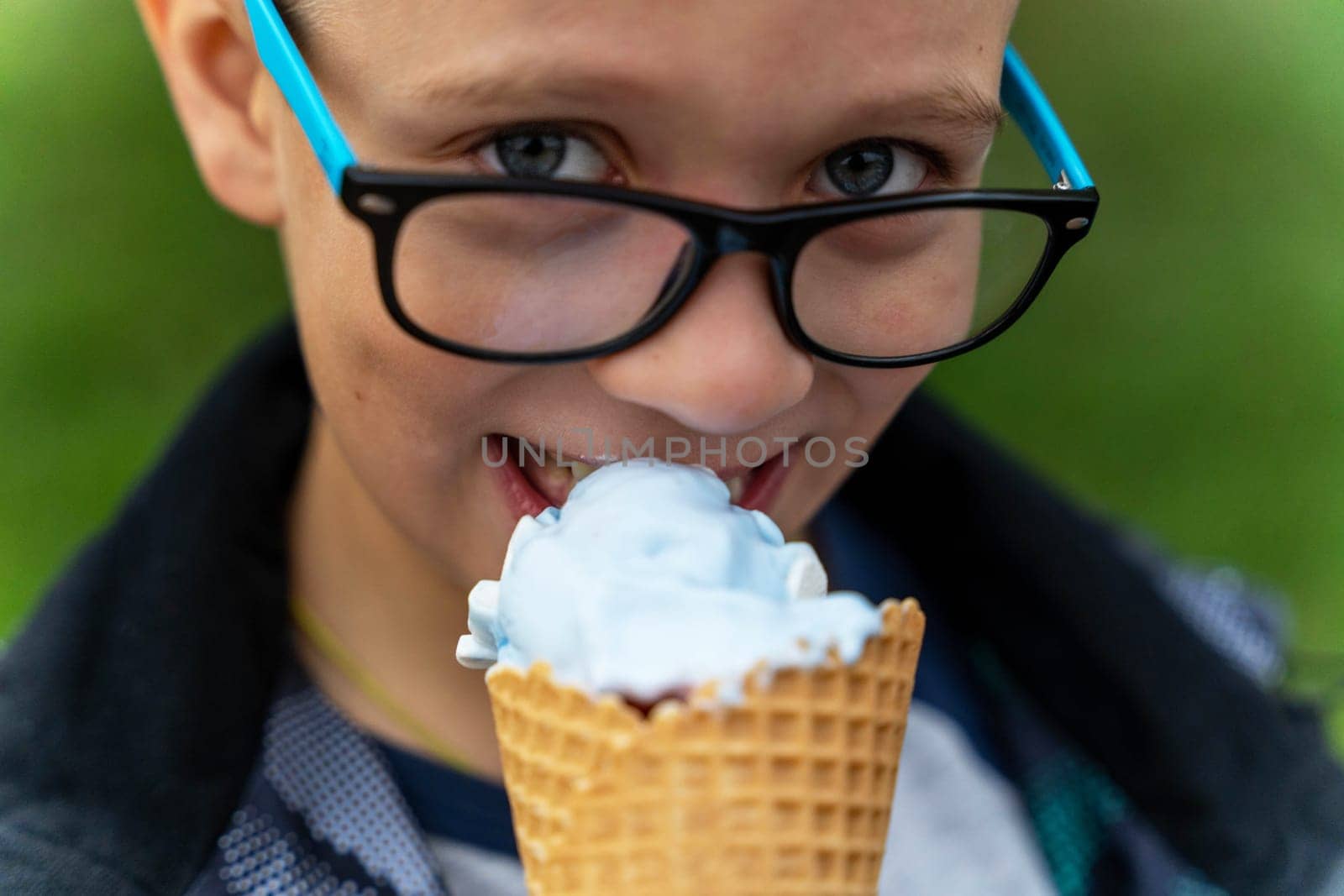 Funny Caucasian boy eats ice cream in a waffle cone on the street in the park, smiles and looks at the camera. A happy Caucasian boy with glasses walks in the park.