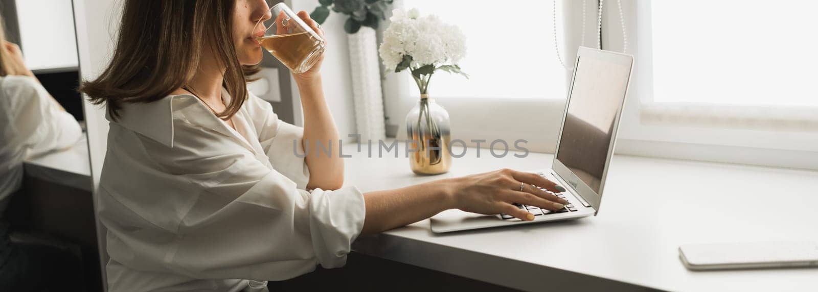 Close-up of young pretty woman drinking tea while break after typing and browsing online on laptop working on computer while sitting in cozy room alone. Distance learning and work with break for coffee time concept