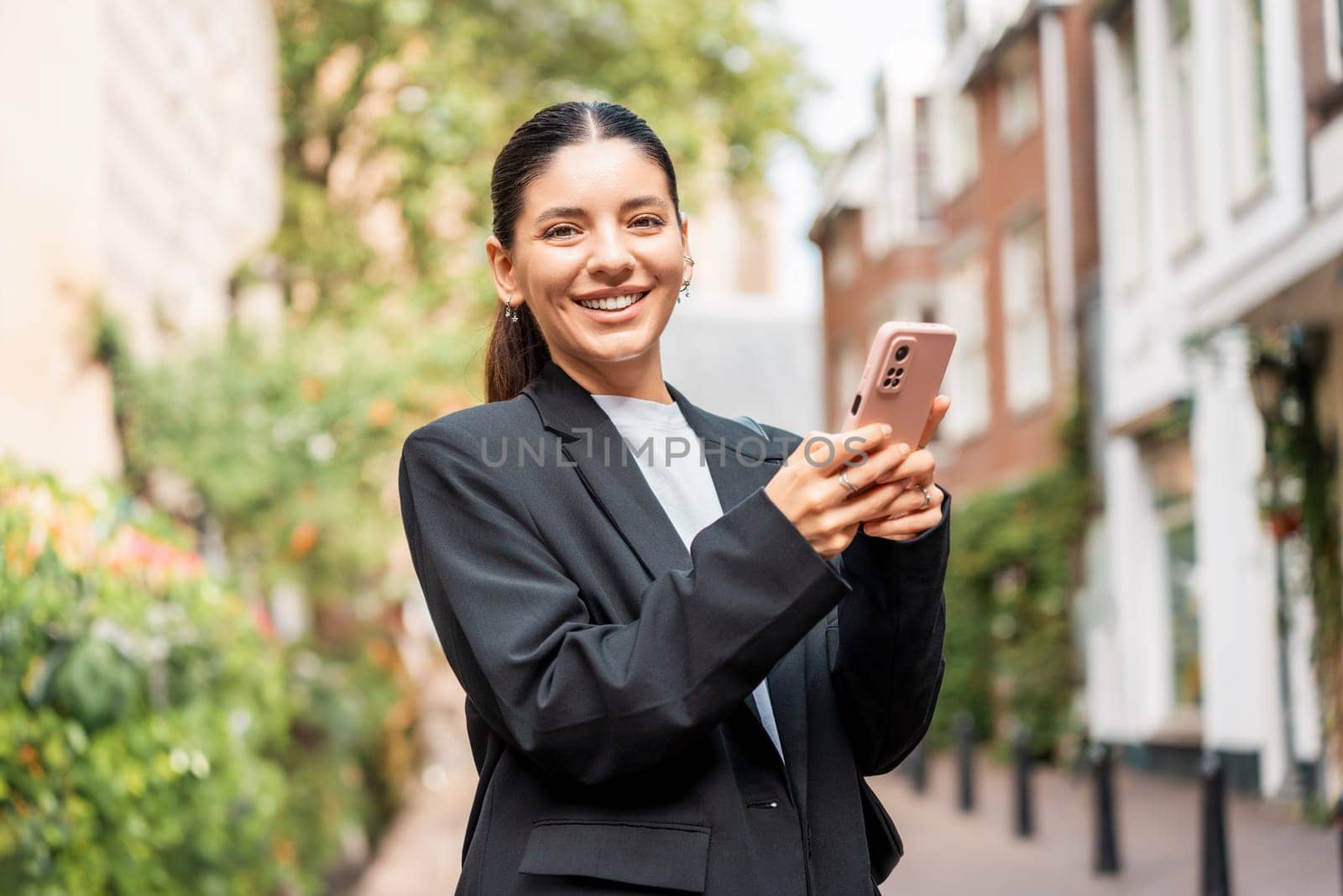 Beautiful confident Hispanic multiracial young businesswoman in black suit and white t-shirt holding a phone in the streets smiling to camera. New entrepreneur by AndreiDavid