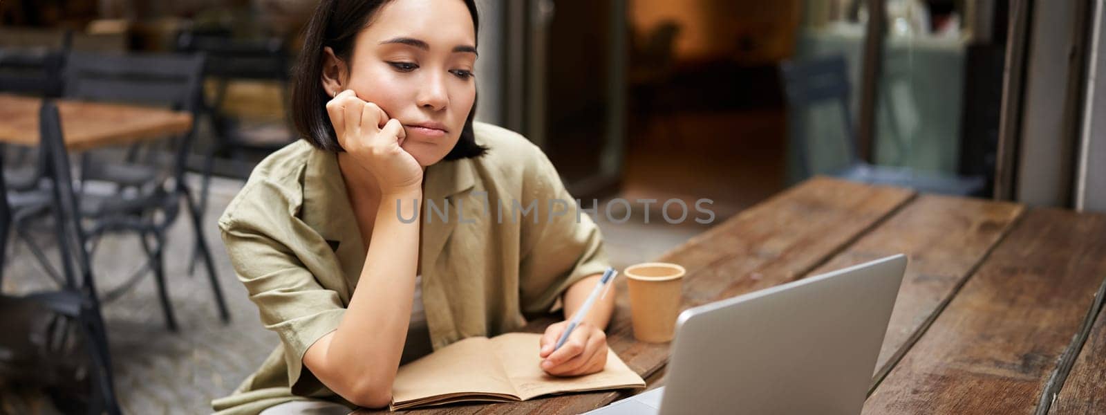 Portrait of young korean girl making notes, listening online meeting, lecture, looking at laptop screen, working remotely from cafe.