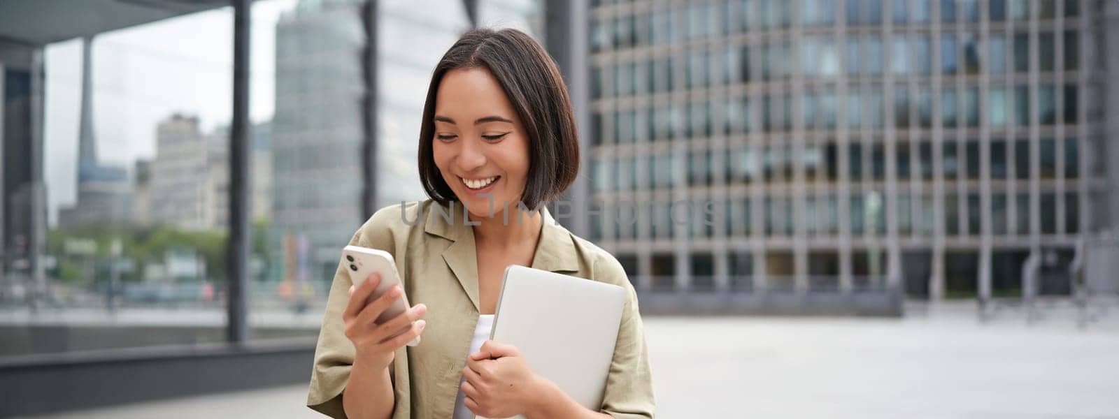 Portrait of young smiling asian woman walking on street, going to work with laptop and smartphone.