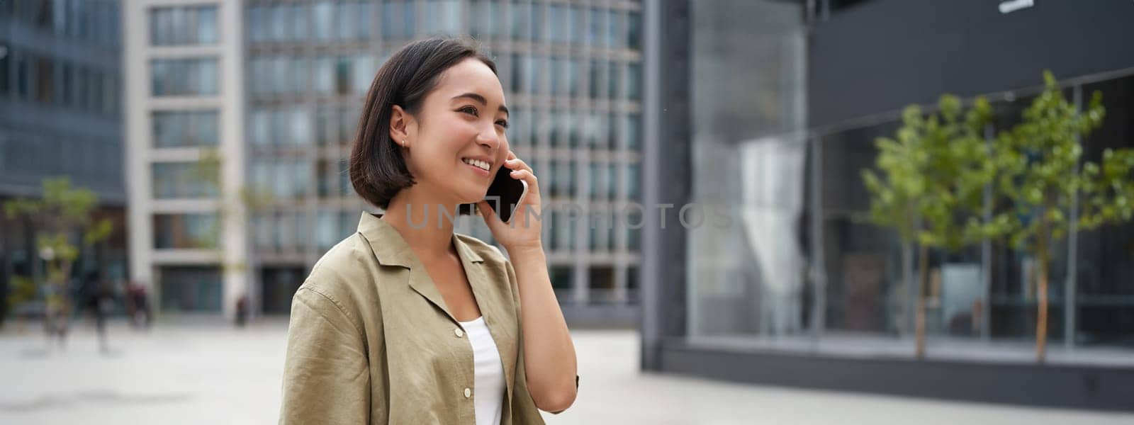 Vertical shot of young asian digital nomad, girl talks on mobile phone and walks on street with laptop. Young woman remote worker going to coworking space by Benzoix