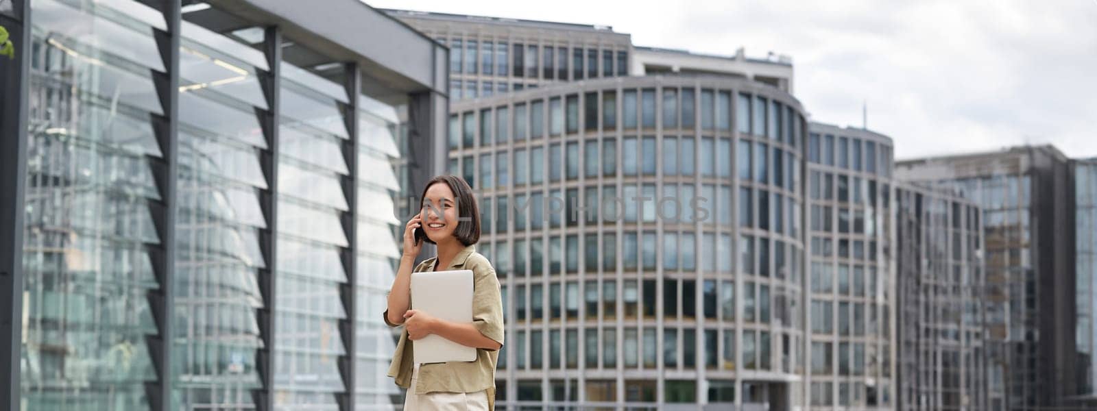 Vertical portrait of stylish asian girl student, talking on mobile phone while walking, holding laptop.