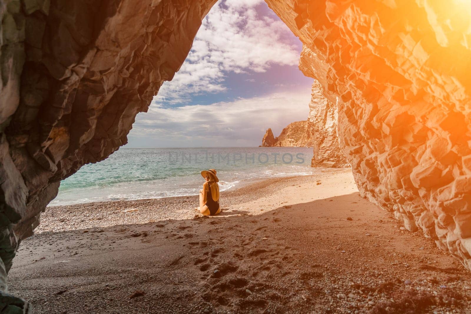 Woman travel sea. View of a woman in a black swimsuit from a sea cave Attractive woman enjoying the sea air sits on the beach and looks at the sea. Behind her are rocks and the sea by Matiunina