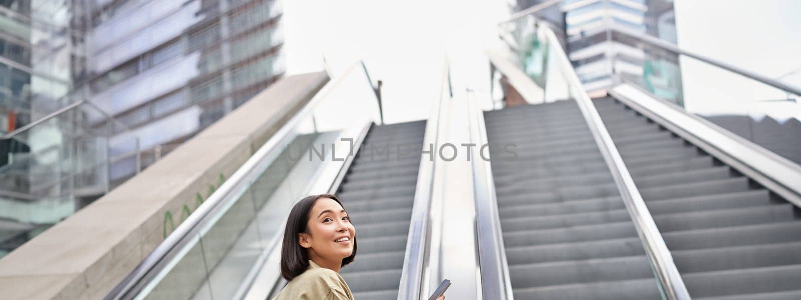 Young happy woman standing on escalator with smartphone, going up, walking in city by Benzoix