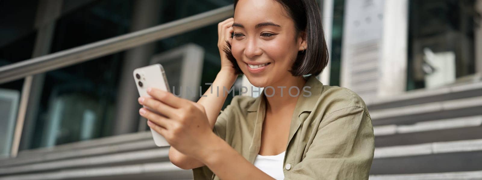 Portrait of asian girl takes selfie on mobile phone. Korean woman smiling, video chat on smartphone app while sits outside on stairs by Benzoix