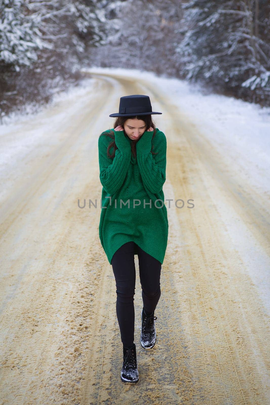 A young girl in a green sweater stands in the middle of a snowy road in a thick pine forest. Freezing day
