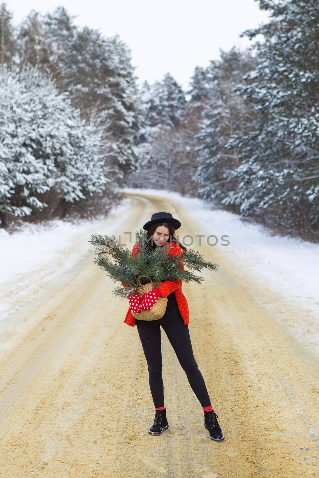 A girl in a red sweater and hat, in red mittens holds a basket with pine branches in her hands stands in the middle of a snow-covered road in a forest with pine branches