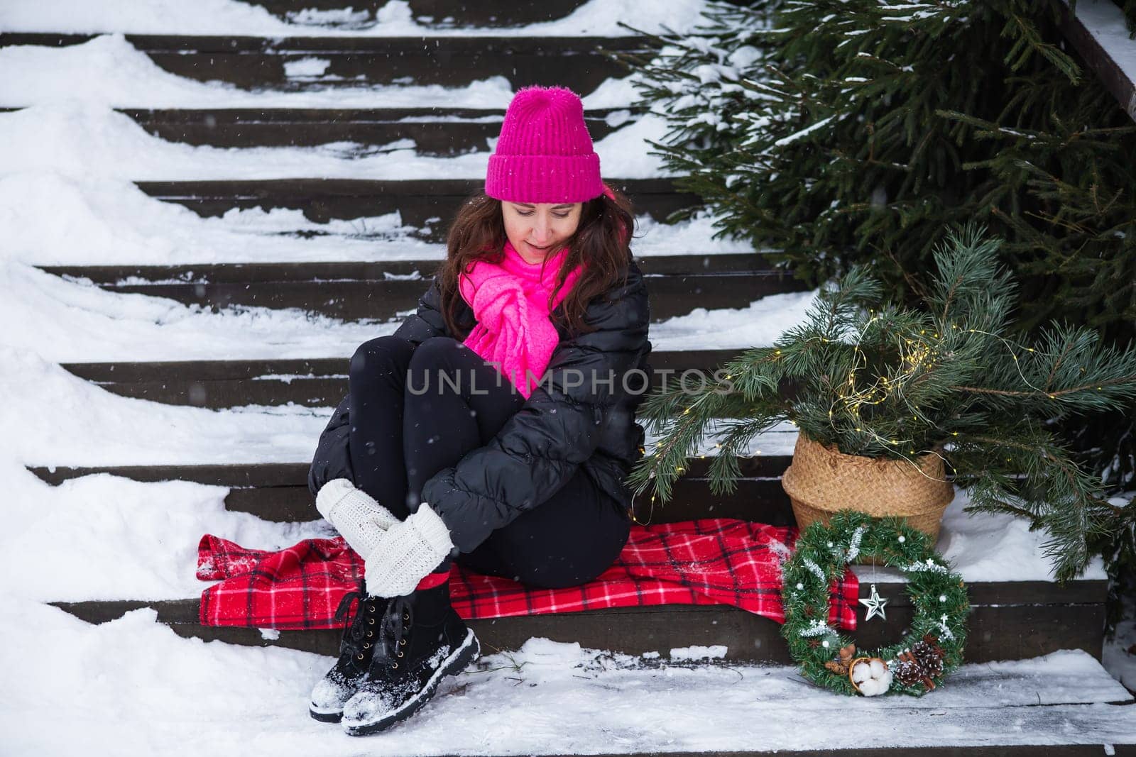 A woman sits on wooden stairs covered with snow in a pink hat near a Christmas tree in a basket, lights, gifts. New Year's magical atmosphere of coziness, love and warmth