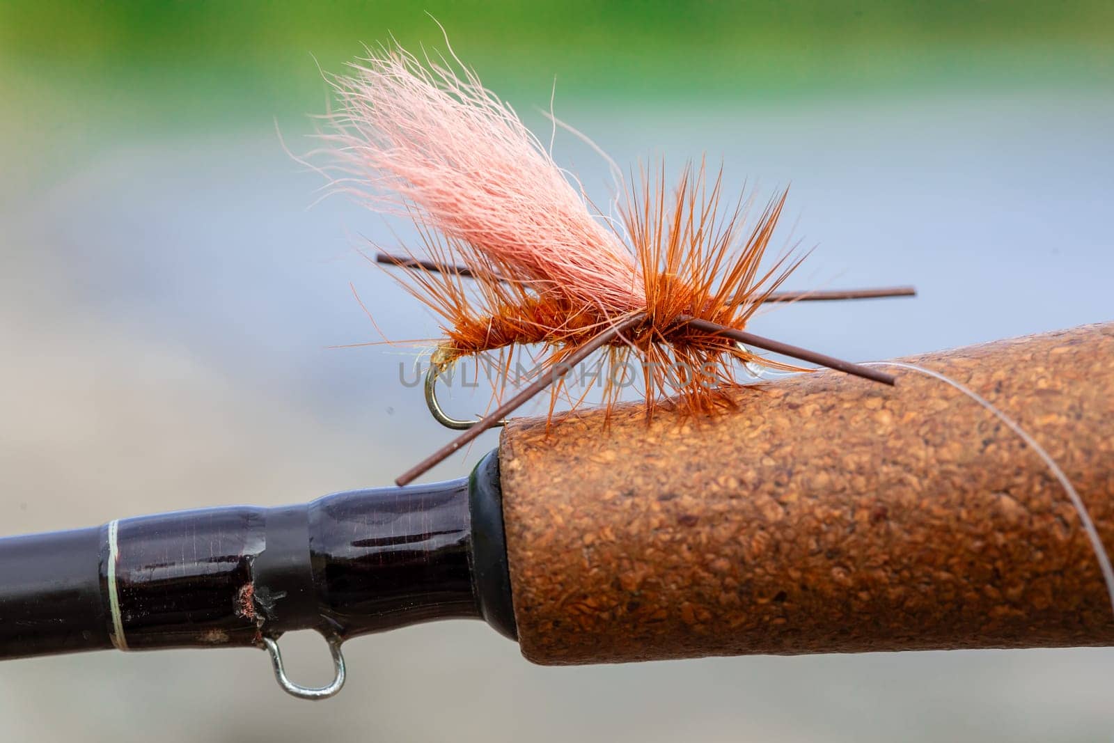 Dry fly pattern in orange with rubber legs on the cork grip of a fly fishing rod at the river.