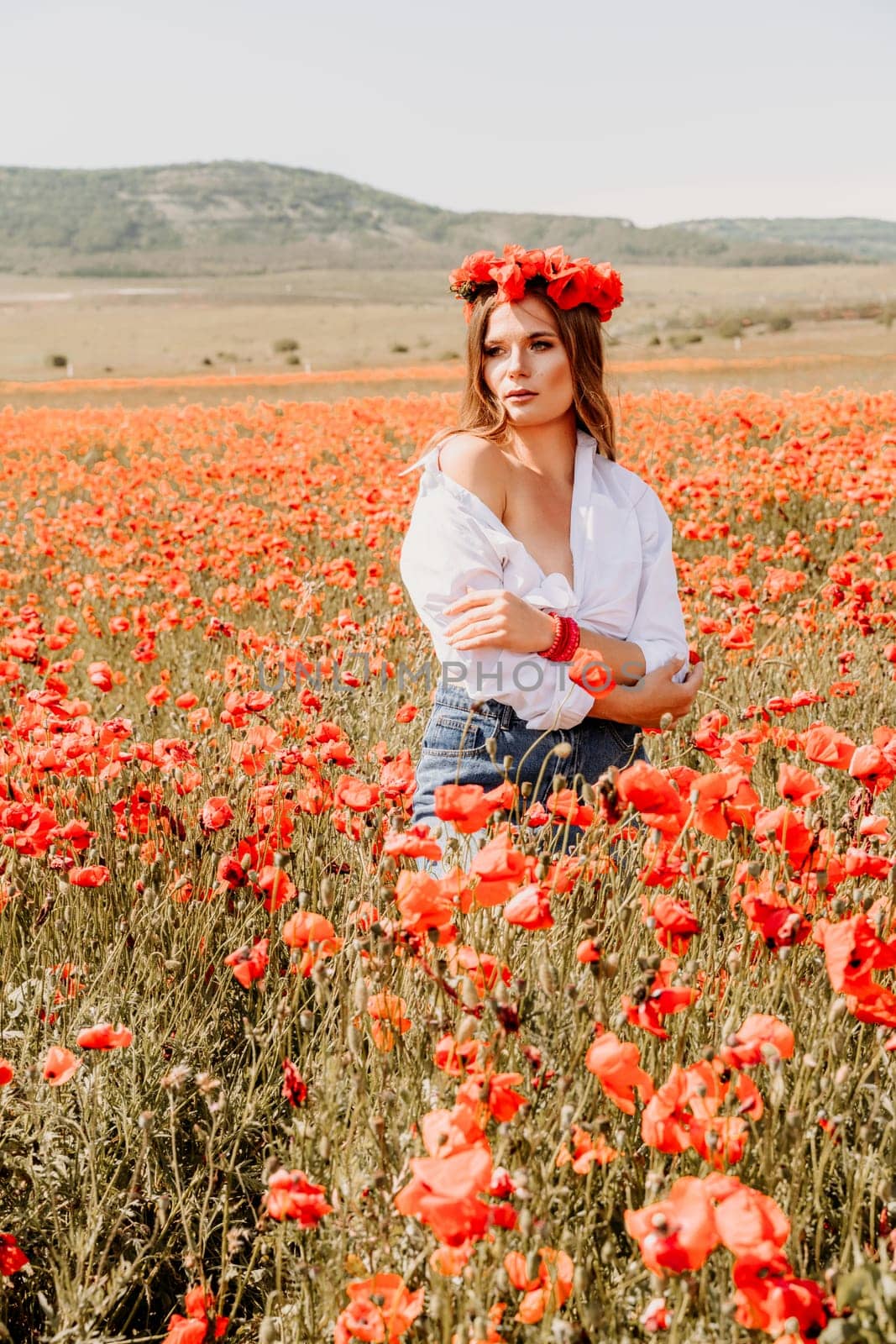 Happy woman in a poppy field in a white shirt and denim skirt with a wreath of poppies on her head posing and enjoying the poppy field. by Matiunina