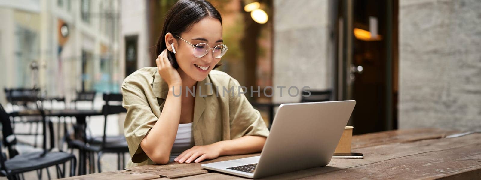 Portrait of young woman video chatting, having online meeting on laptop, sitting in outdoor cafe with compute, wearing glasses and smiling.