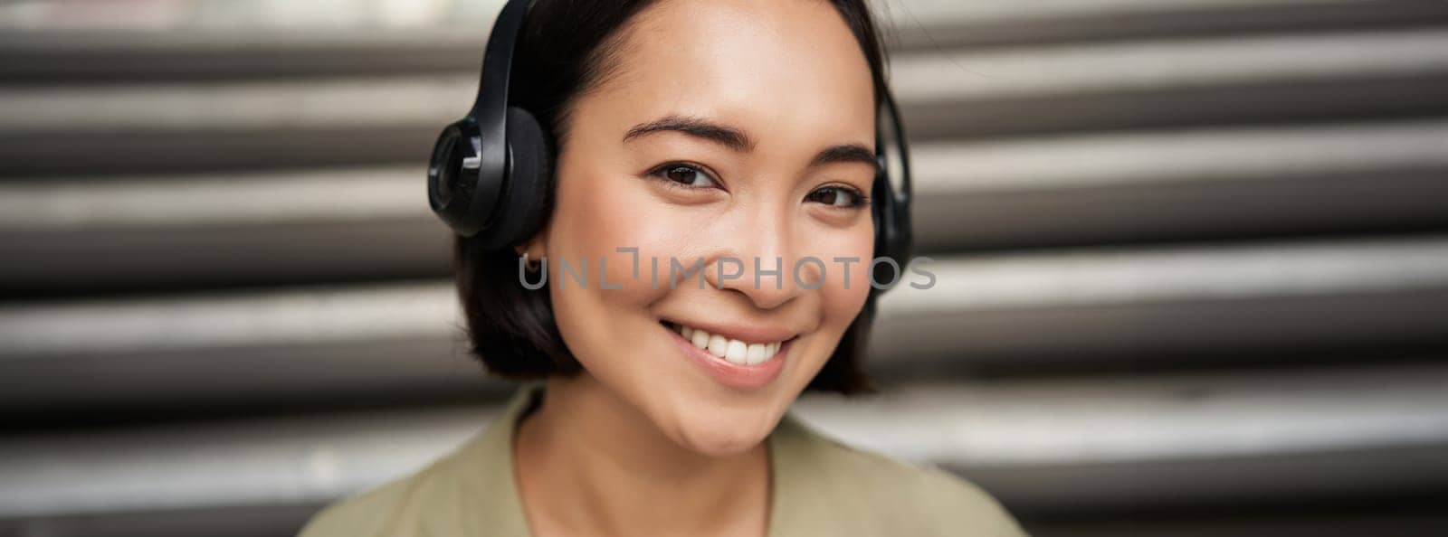 Close up portrait of smiling asian girl in headphones, listens to music outdoors, looking happy. People concept