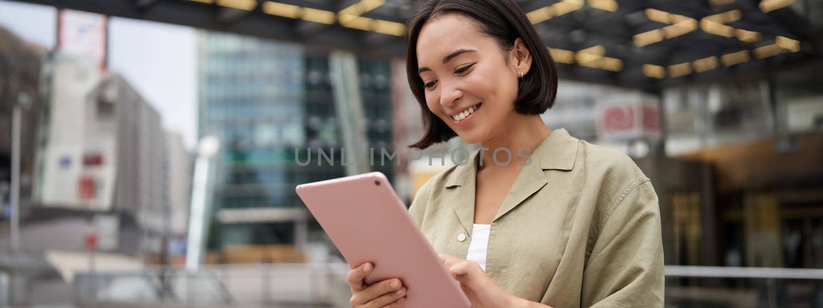 Portrait of asian woman standing on street, using tablet, smiling with carefree face expression, outdoor shot by Benzoix