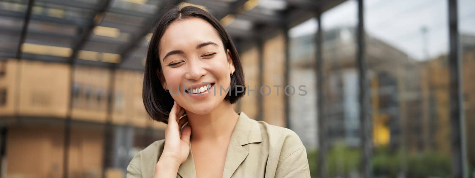 Close up portrait of asian girl with happy face, smiling and laughing, touching her skin, standing on street on sunny day by Benzoix