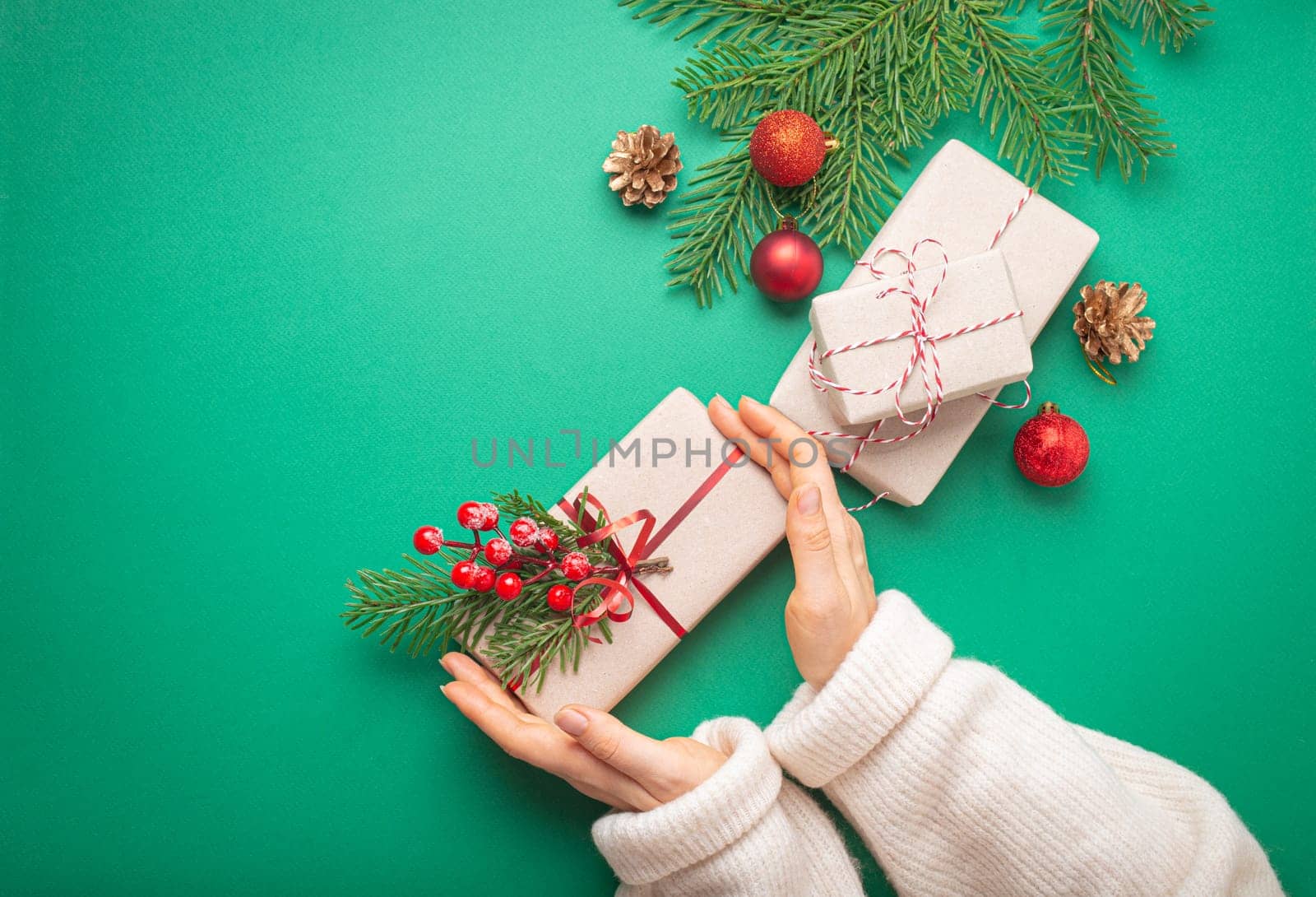 Christmas or New Year celebration green paper festive background with female hands holding wrapped present box, decoration fir tree, cones, berries, sparkly red balls. Space for text. by its_al_dente