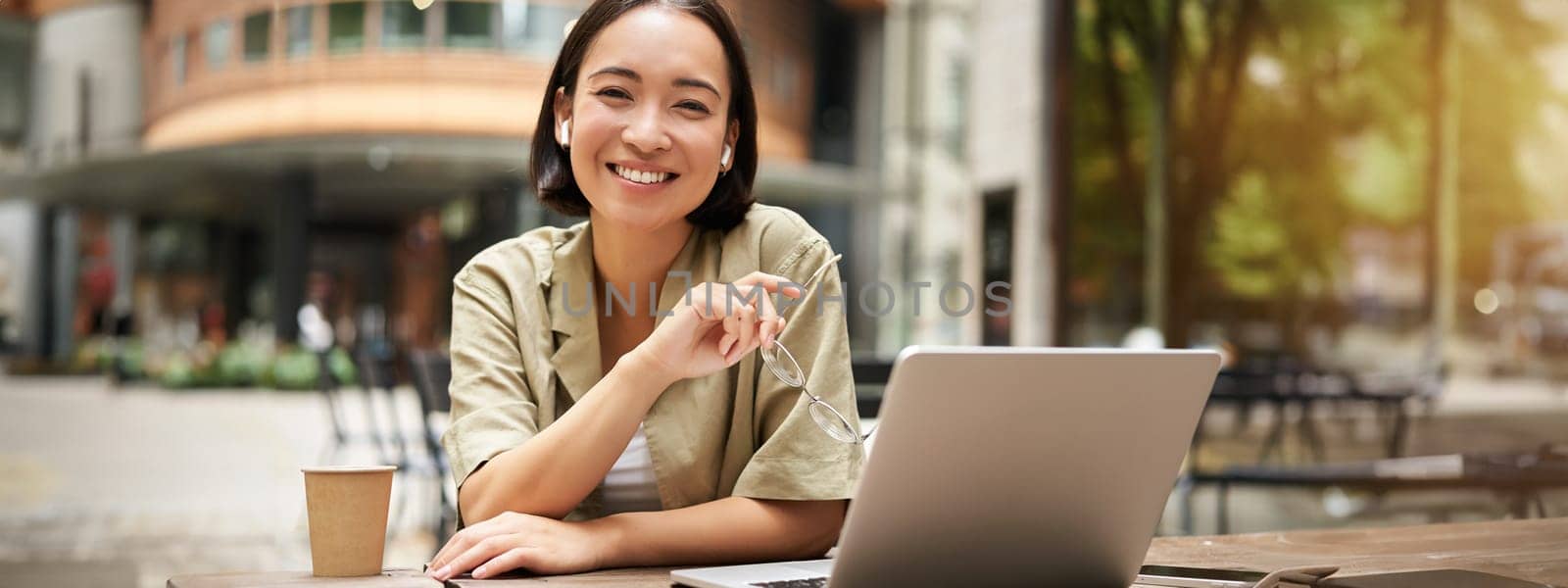 Urban people. Young woman working with laptop from cafe, coworking space, sitting outdoors with cup of coffee and smiling at camera, holding glasses.