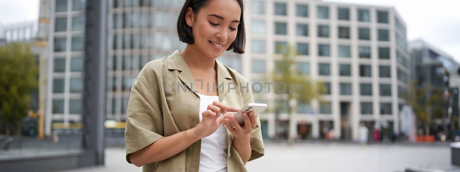 Mobile technology. Smiling asian woman using smartphone app, looking at her telephone on street, checking map, calling or texting someone.