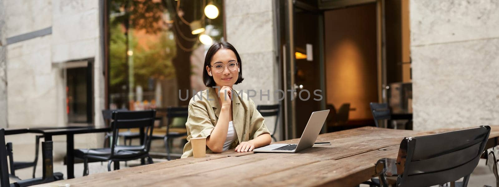 Portrait of asian girl in glasses, sitting with laptop in cafe, working remotely, studying online, posing with coffee and computer.