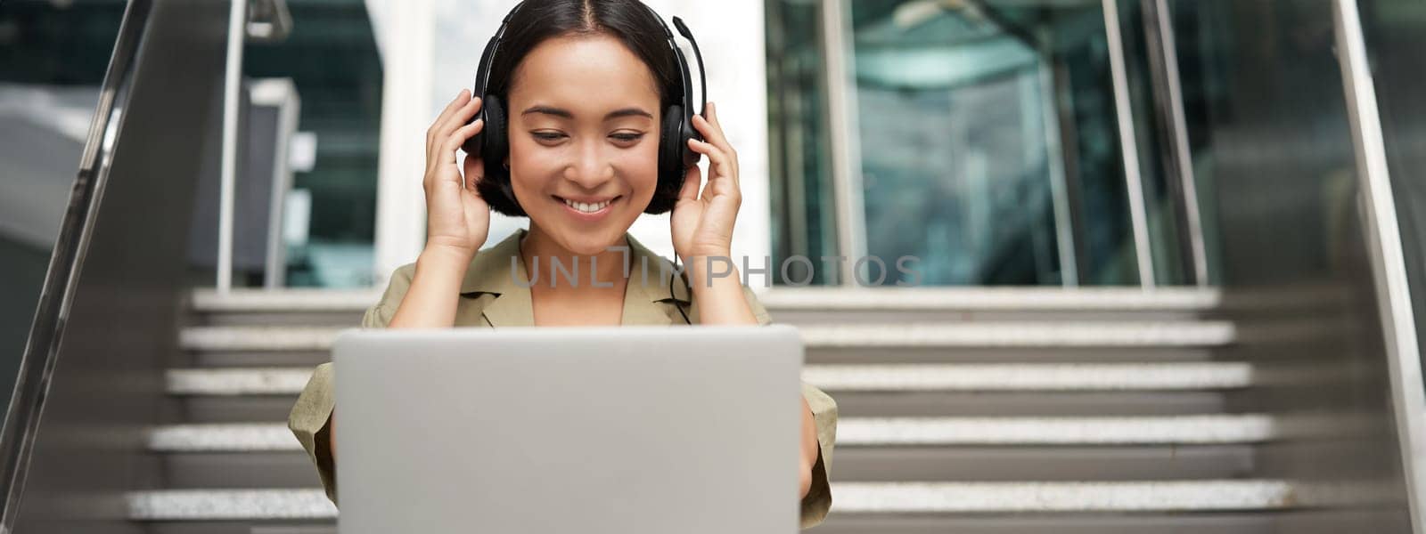 Portrait of young asian woman sitting with laptop and headphones, watching video, does online course on computer, sitting on stairs outdoors by Benzoix