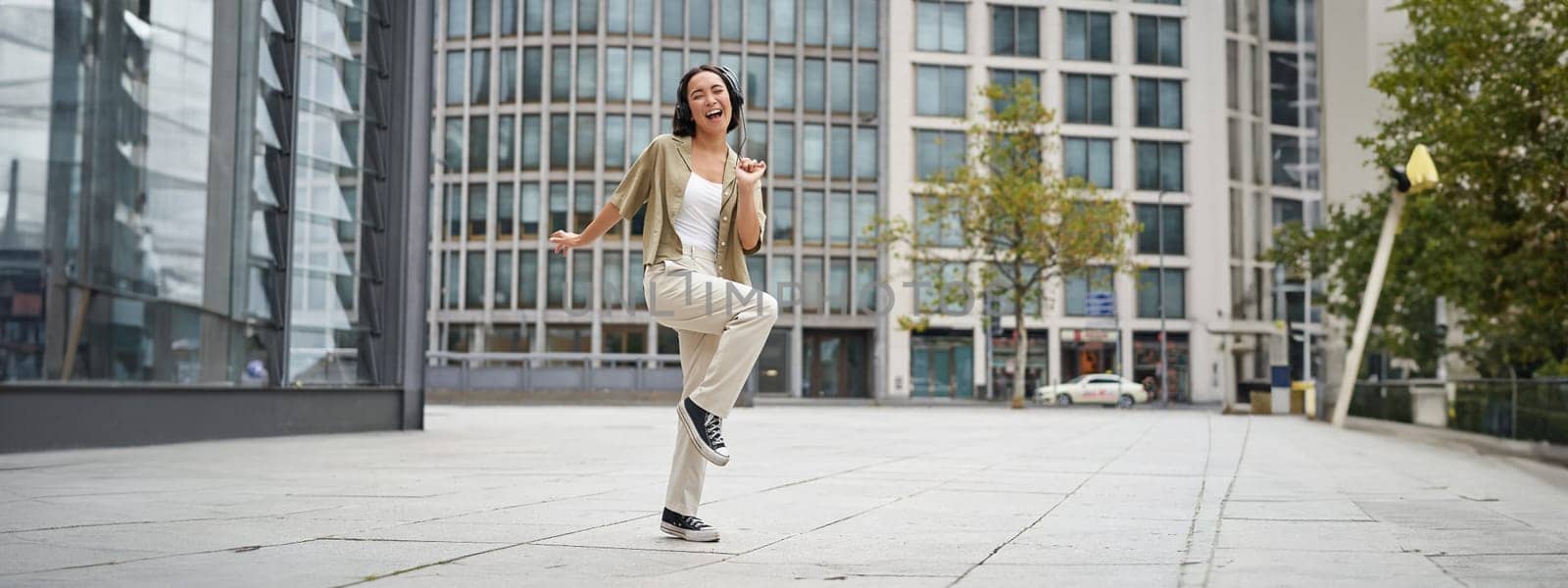 Happy people in city. Upbeat young girl dancing on street in headphones, listening music in headphones by Benzoix