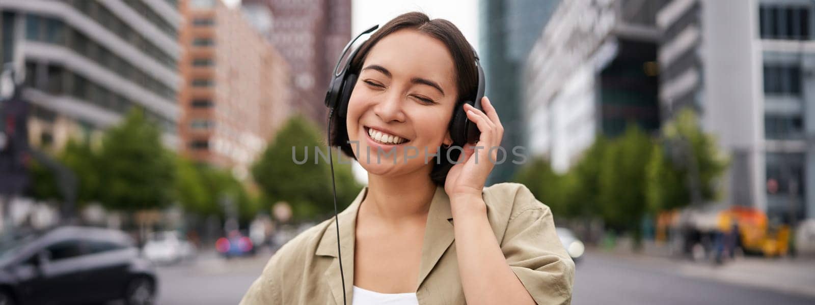 Portrait of smiling asian woman in headphones, standing in city centre on street, looking happy, listening to music.