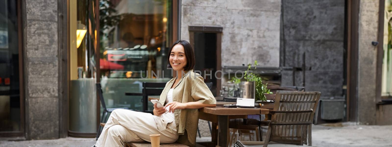 Portrait of stylish young korean woman sits in cafe, holds smartphone, smiles, enjoys coffee outdoors by Benzoix