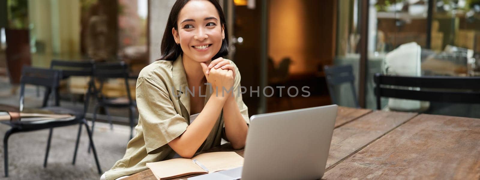 Portrait of asian girl works outdoors in cafe, sits with laptop, studies, smiles happy.