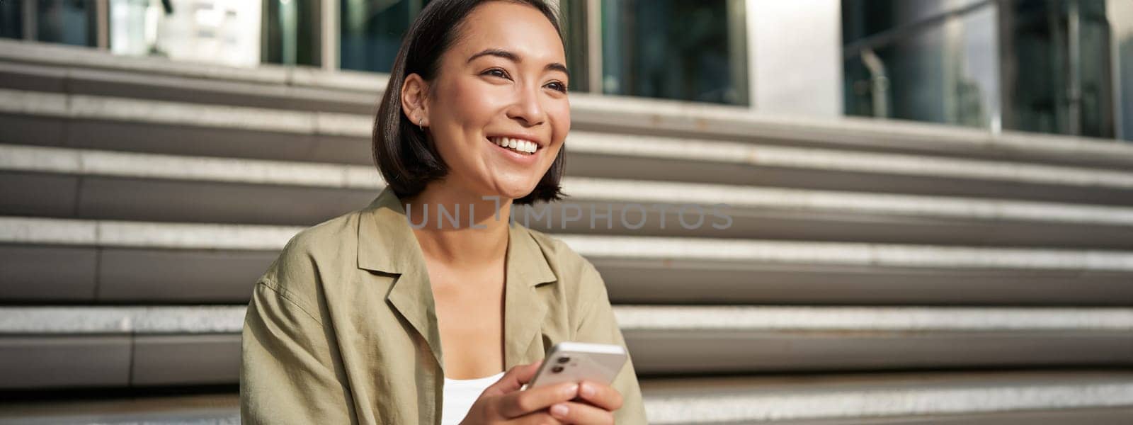 Beautiful asian girl sits on stairs with smartphone. Young korean woman resting outdoors, using mobile phone application by Benzoix