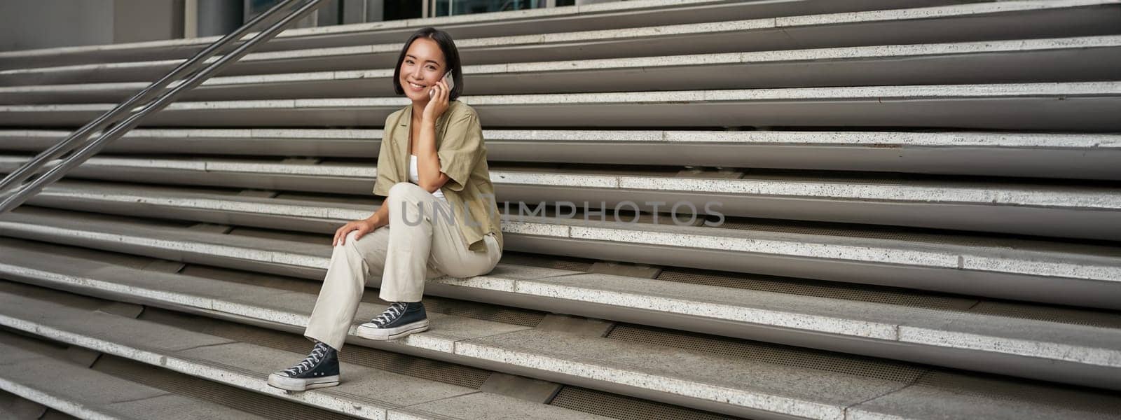 Smiling asian girl sits on stairs of building and talks on mobile phone, relaxing during telephone conversation by Benzoix