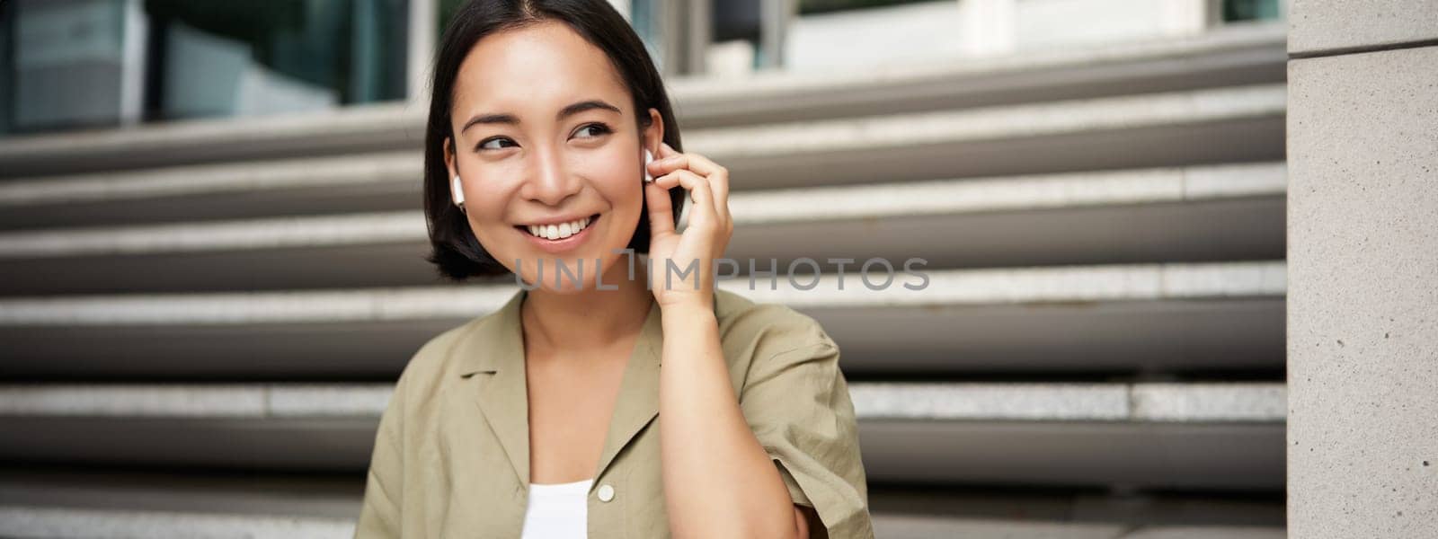 Close up portrait, street photo of asian woman listening music in wireless headphones, smiling, sitting on stairs.
