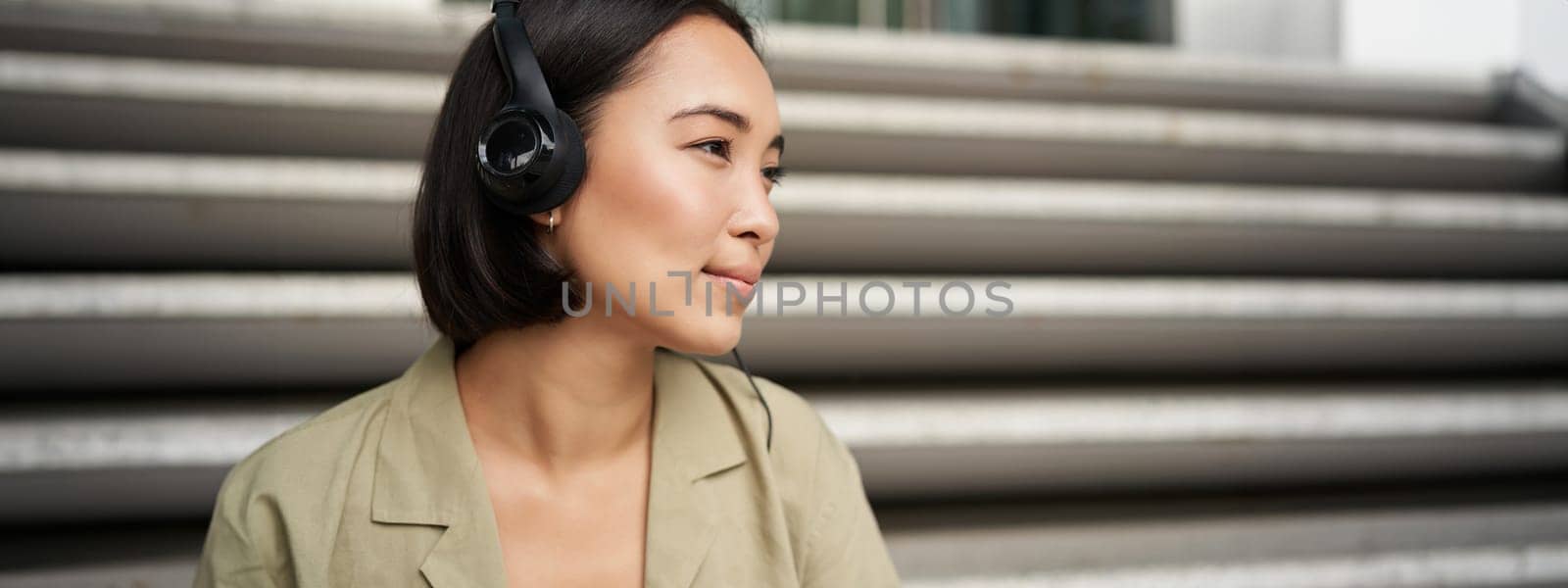 Young asian woman listening to music in headphones, sitting outside on street, smiling by Benzoix