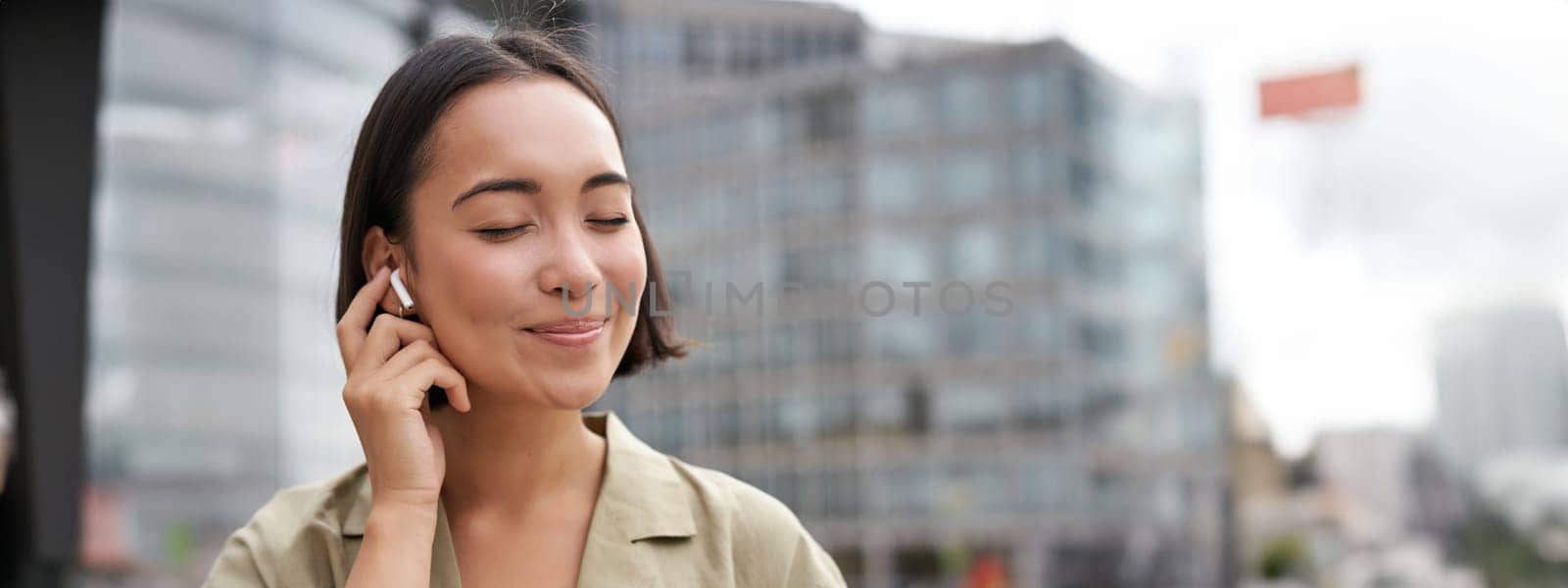 Vertical shot of smiling asian woman in wireless headphones, enjoys listening to music in earphones, holds mobile phone by Benzoix