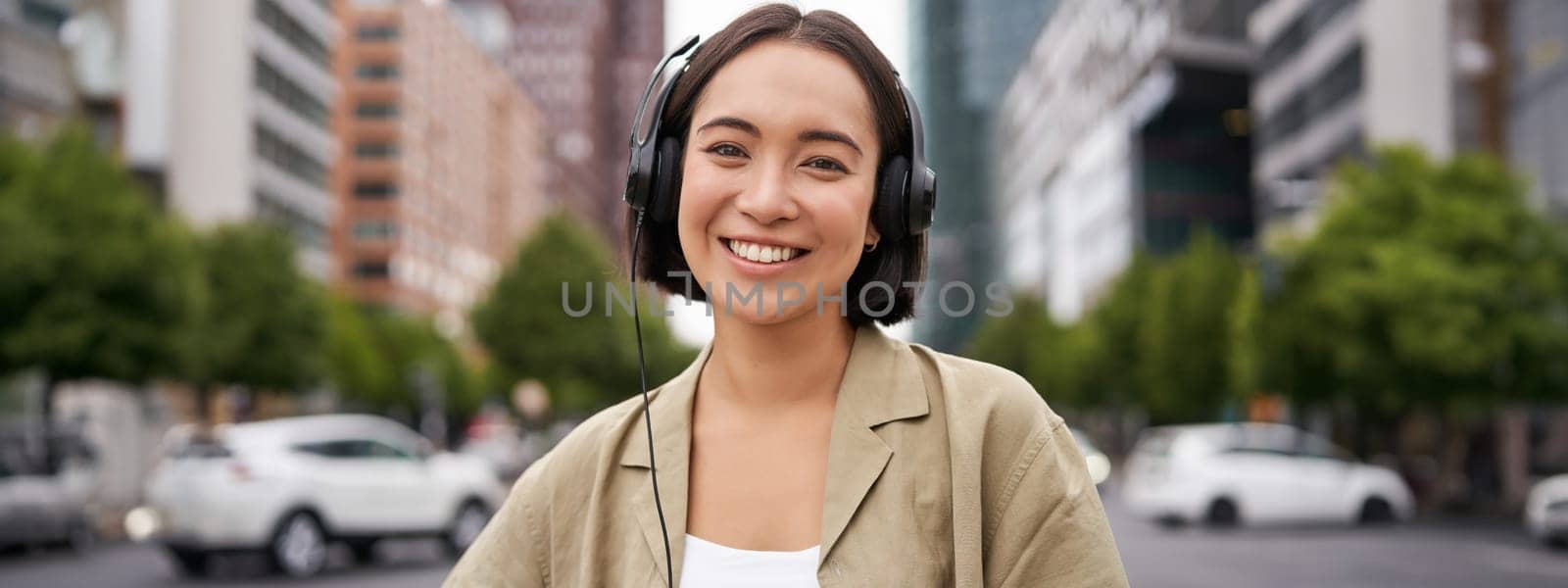Portrait of smiling asian woman in headphones, standing in city centre on street, looking happy, listening to music by Benzoix