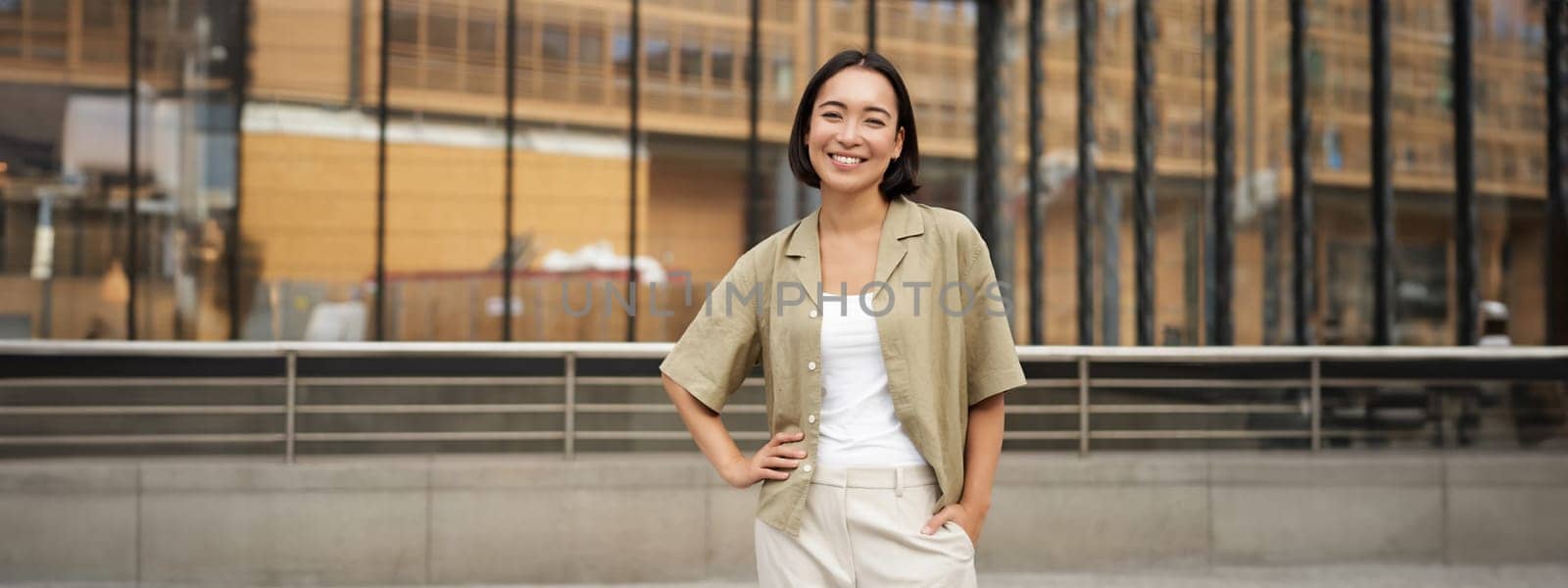 Portrait of young confident female model, girl in casual clothes, posing outside near glass building, smiling at camera.