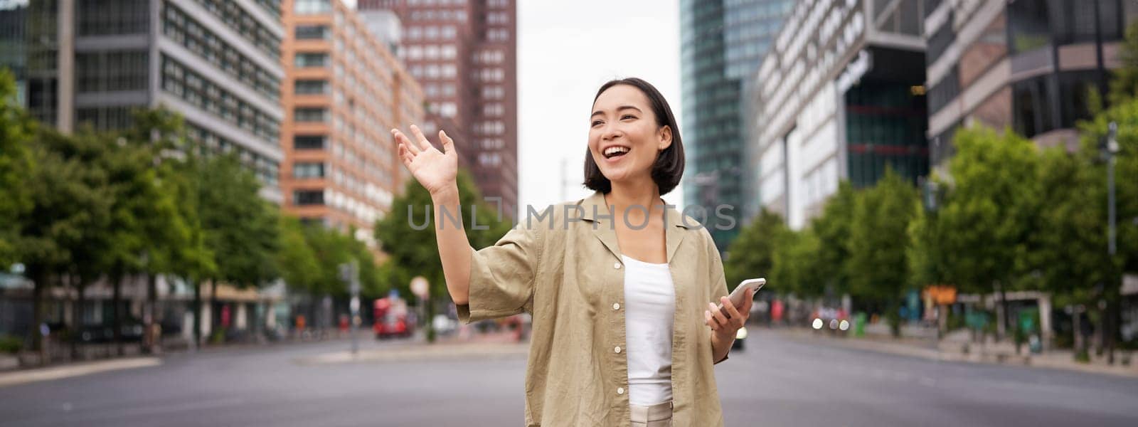 Happy asian girl passing by friend and waving at them on street, saying hello while walking in city, holding smartphone by Benzoix