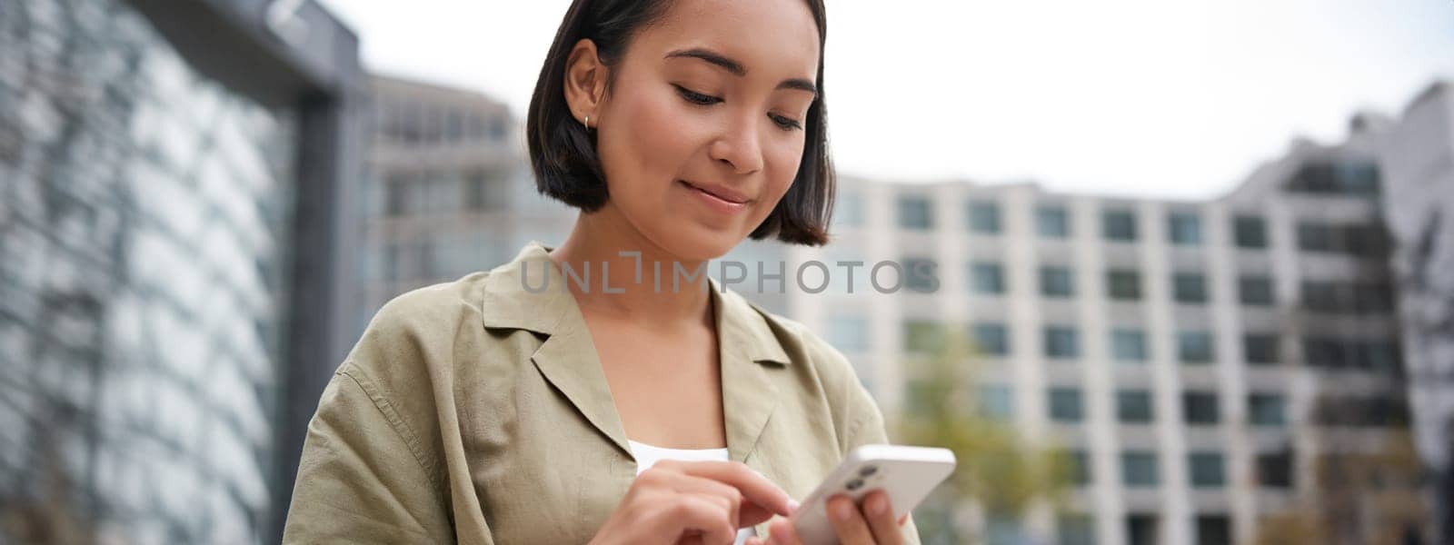 Mobile technology. Smiling asian woman using smartphone app, looking at her telephone on street, checking map, calling or texting someone by Benzoix